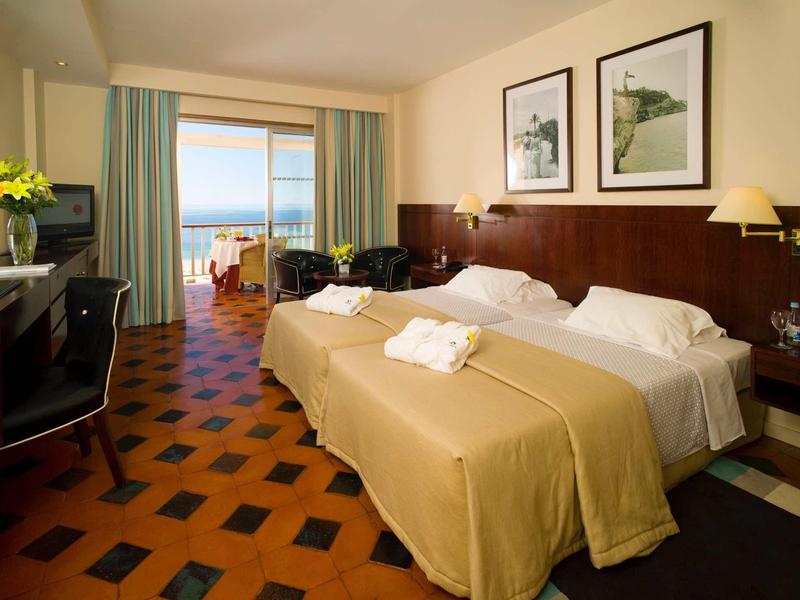 Chambre d'hôtel lumineuse avec deux lits, balcon donnant sur la mer et fleurs fraîches.