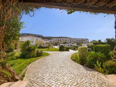 Pflasterweg führt durch Garten zu weißen Gebäuden unter blauem Himmel.