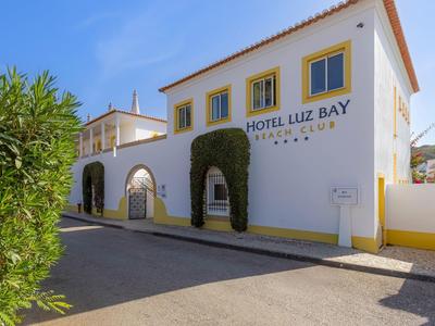 Außenansicht des Hotels Luz Bay mit weißer Fassade und gelben Akzenten unter klarem Himmel.