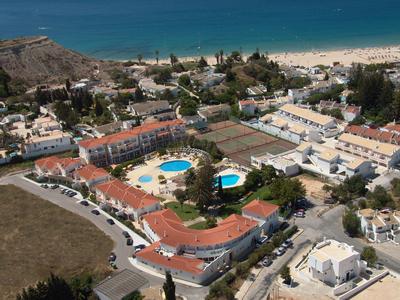 Luftaufnahme eines Ferienorts mit mehreren weißen Gebäuden, zwei Pools und einem Strand im Hintergrund.