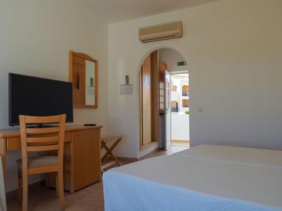 Hell eingerichtetes Schlafzimmer mit Doppelbett, Holzmöbeln, Fernseher und offener Tür zum Flur.