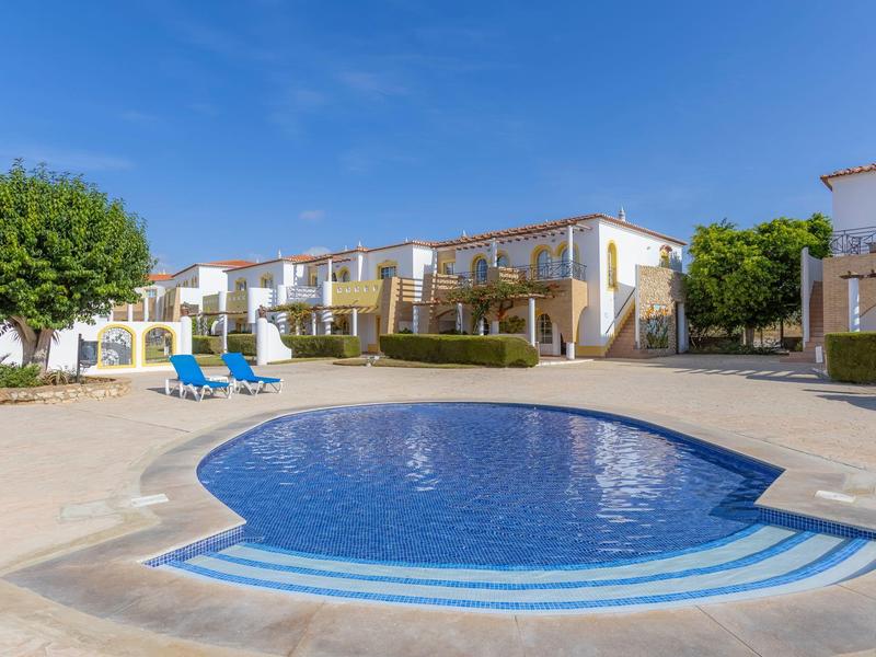 Rundes Schwimmbecken vor Reihenhäusern mit blauen Liegestühlen und Bäumen unter klarem Himmel.