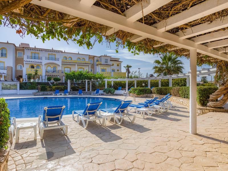 Sonniger Poolbereich mit blauen Liegestühlen und gepflegtem Garten vor Hotelgebäuden.