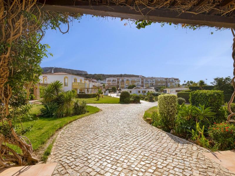 Pflasterweg führt durch Garten zu weißen Gebäuden unter blauem Himmel.
