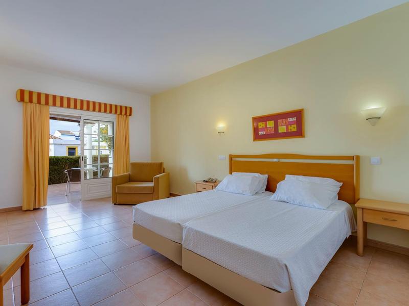 Helles Hotelzimmer mit zwei Betten, Holzmöbeln und Zugang zu einer Terrasse mit Blick nach draußen.