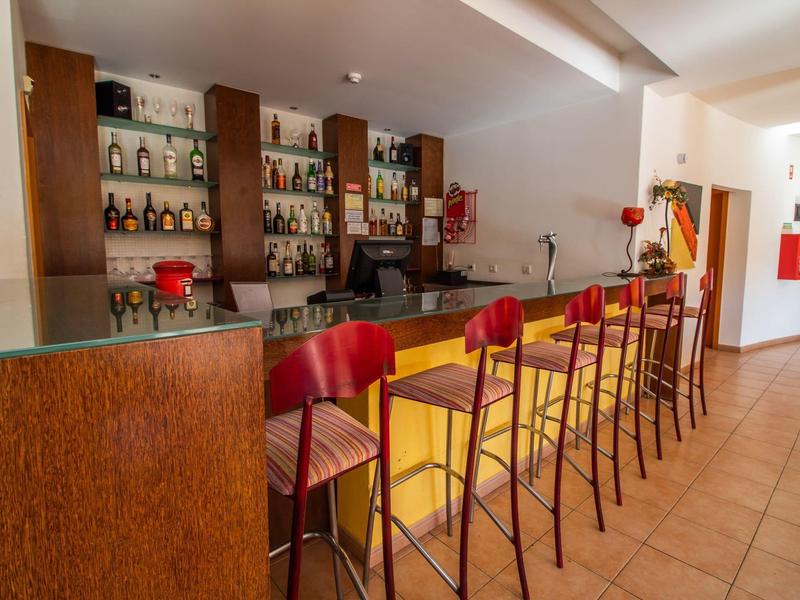 Bar mit Holztheke, rundem Glas, roten Hockern und Regalen mit Flaschen an der Wand.