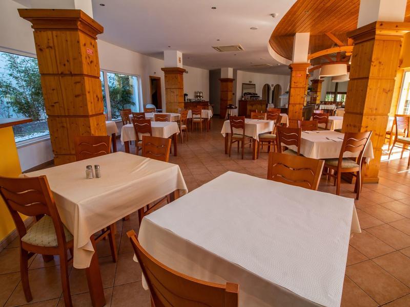 Restaurant mit Holzmöbeln, quadratischen Tischen und großen Fenstern, warme Lichtstimmung.