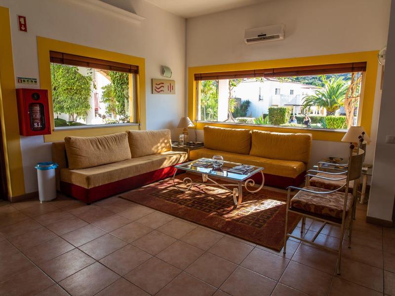 Hell eingerichteter Wohnraum mit gelben Sofas, großen Fenstern und braunem Teppich auf Fliesenboden.