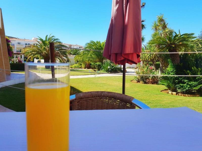 Glas mit Orangensaft auf weißem Tisch, im Hintergrund Garten mit grünem Rasen und rotem Sonnenschirm.