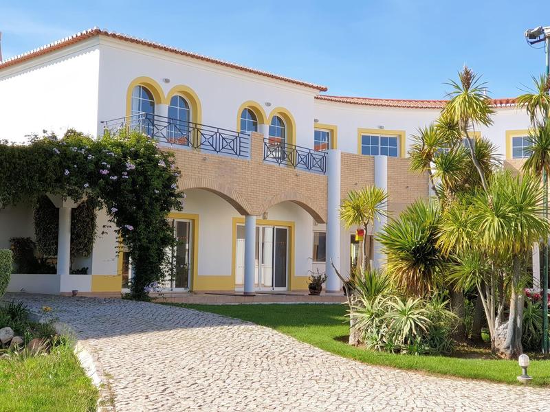 Großes weißes Haus mit gelben Akzenten, Rundbögen, grünem Garten und Palmen davor.