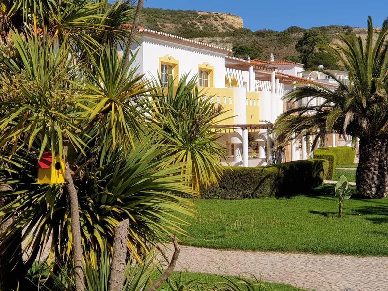 Weiße Villa mit gelben Balkonen, umgeben von grünen Palmen und Rasen vor blauem Himmel.