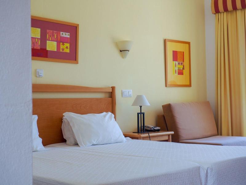 Zweibettzimmer mit weißen Betten, Holzkopfteil, seitlichem Nachttisch, Sofa und gelben Vorhängen.