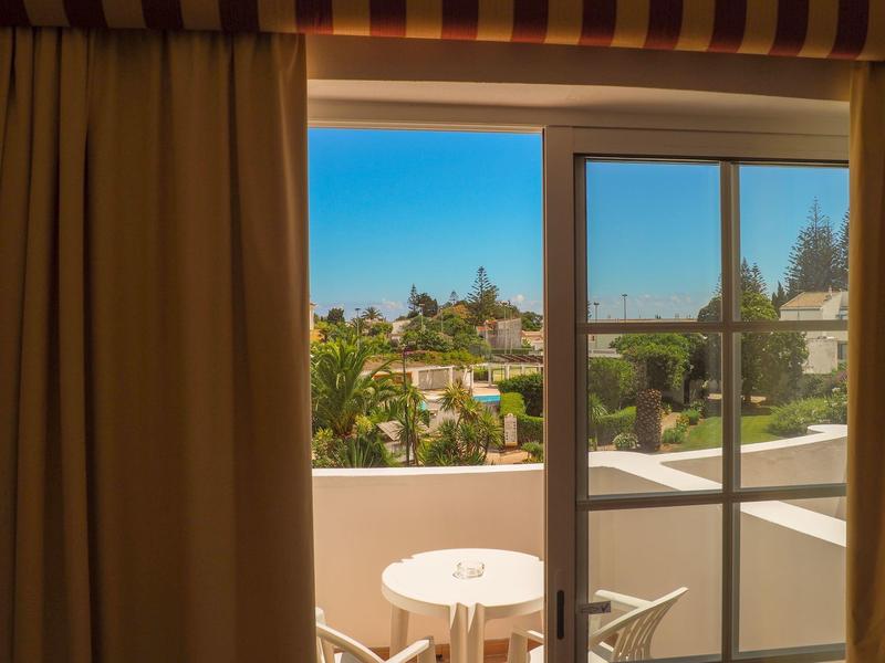 Balkon mit weißem Geländer, kleinem runden Tisch und Stuhl, Blick auf Häuser und blauen Himmel.