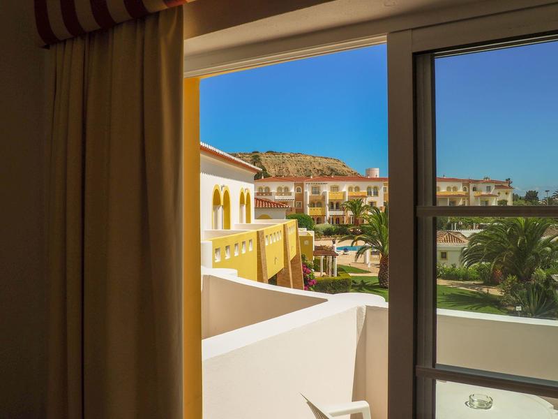 Blick aus Fenster auf sonnige Häuser, grüne Palmen und blauen Himmel hinter braunem Hügel.