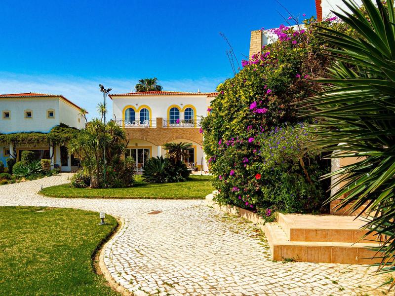 Großes Haus mit gelber Fassade, blauen Fenstern, blühenden Büschen, Kiesweg und grüner Rasenfläche.