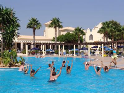 Guests in a hotel pool participating in water activities under palm trees and umbrellas