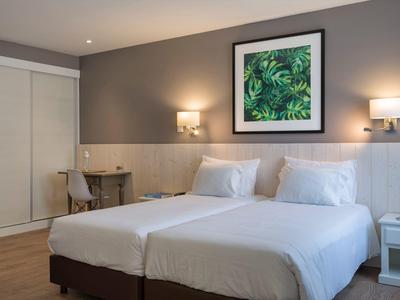 Camera d'hotel moderna con due letti, lampade a muro, scrivania e opera d'arte con piante verdi.