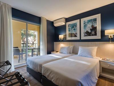 Camera d'hotel moderna con due letti singoli, tonalità blu e balcone con vista su palme.