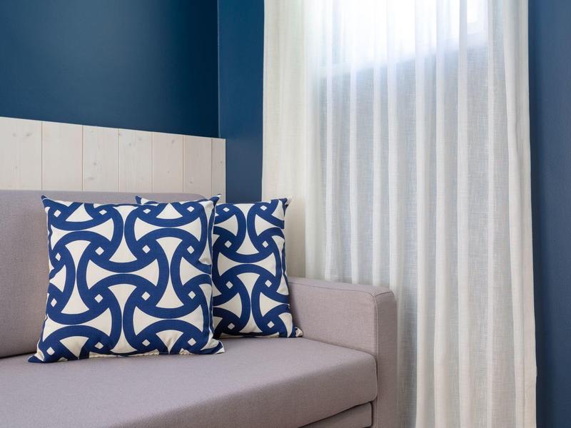 Divano beige con due cuscini blu decorati davanti a una parete blu e tende bianche.