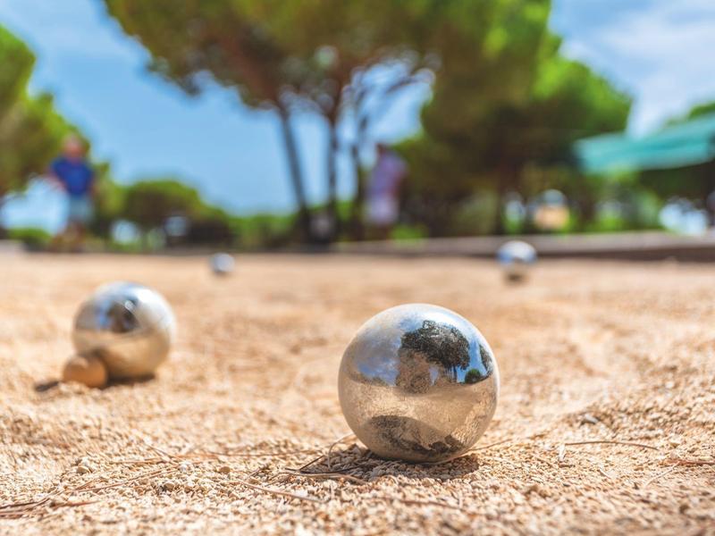 Palline argentate da pétanque giacciono su terreno sabbioso con alberi sullo sfondo.