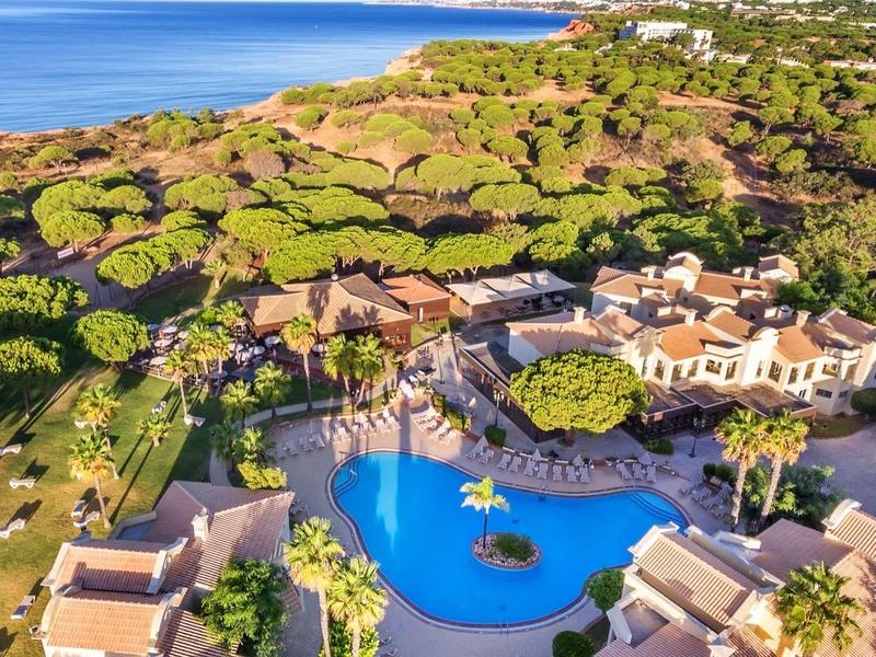 Complesso alberghiero di lusso con grande piscina, circondato da alberi verdi e vicino al mare.