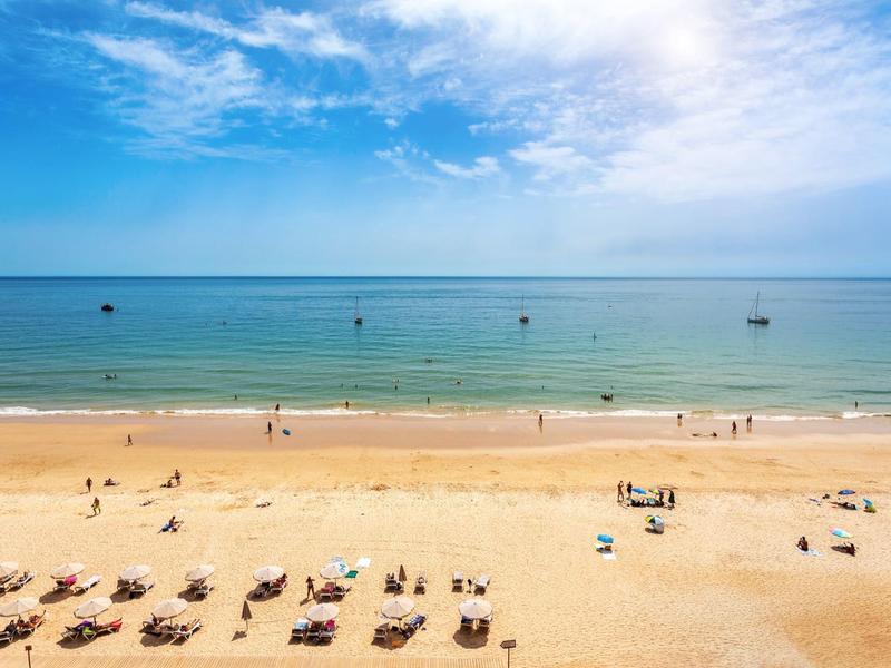 Ampia spiaggia di sabbia con lettini, poche persone e mare calmo sotto un cielo blu.