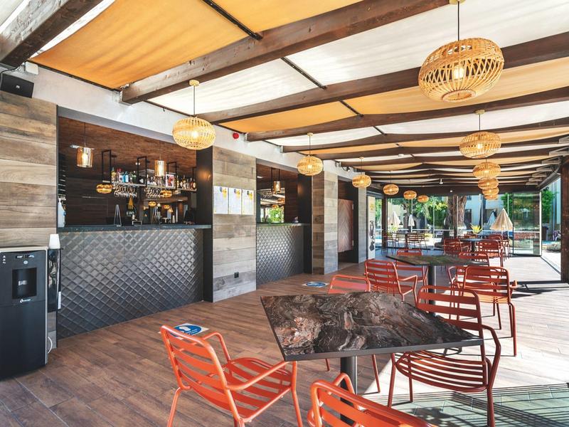 Area bar aperta con sedie arancioni, pavimento in legno e tetto coperto in un hotel.