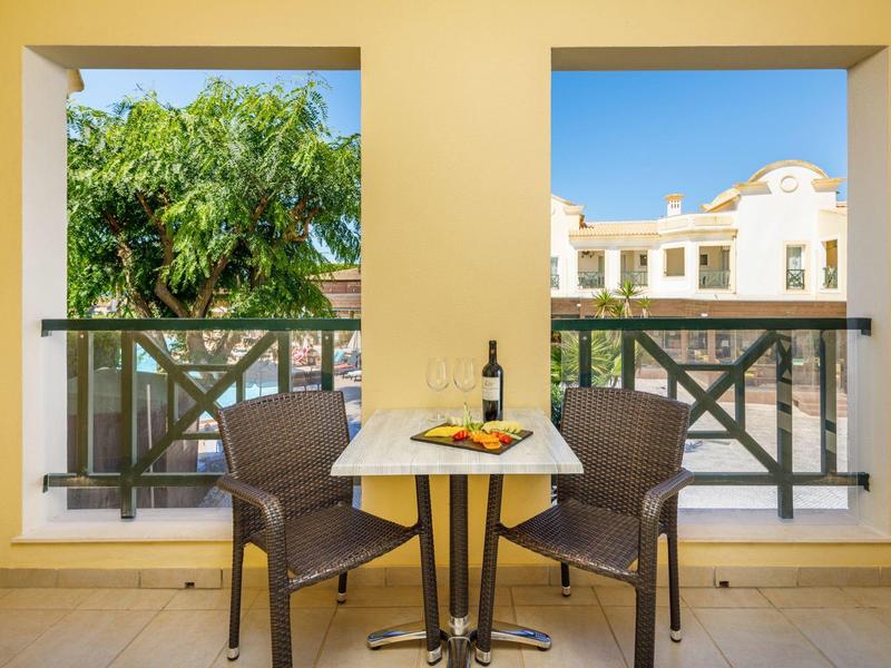 Balcone con tavolo, due sedie, bottiglia di vino, bicchiere e snack davanti a edificio e vegetazione.