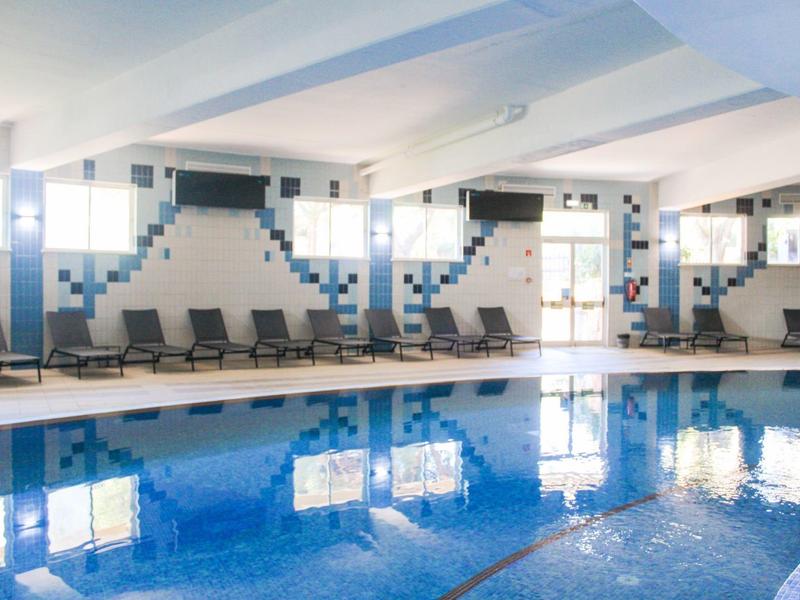 Piscina coperta con piastrelle blu, lettini e grandi finestre lungo la parete.