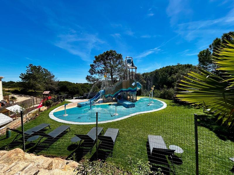Area piscina con scivoli, lettini e prato verde sotto un cielo azzurro.