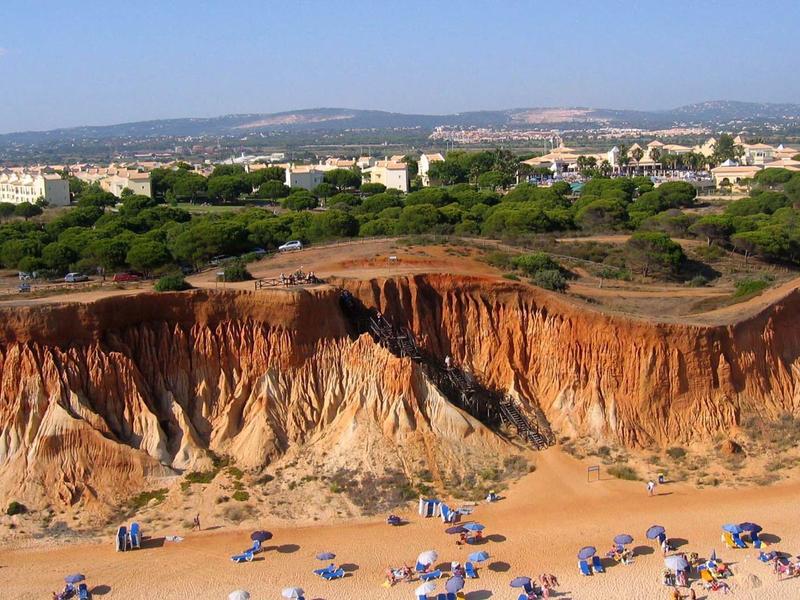 Persone che prendono il sole su una spiaggia sabbiosa sotto alte scogliere rossastre con una città sullo sfondo.