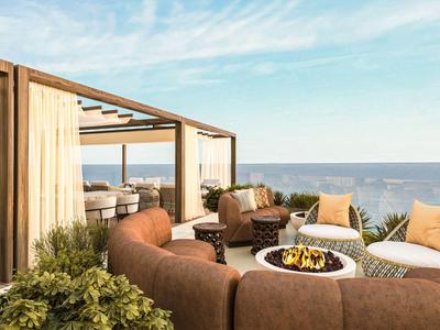 Terrasse eines Hotels mit Sitzgruppen, Feuerstelle und Blick aufs Meer bei klarem Himmel.