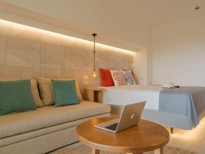 Camera d'hotel moderna con letto matrimoniale, divano e piccolo tavolo rotondo in legno con laptop.