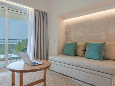 Area di stanza d'hotel moderna con divano, cuscini, tavolo rotondo in legno e balcone con vista sul verde.