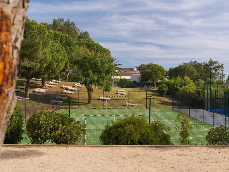 Vista di un campo da tennis recintato circondato da alberi e edifici sotto un cielo soleggiato.