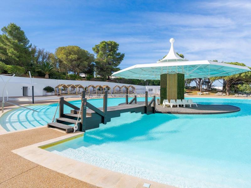 Piscina per il tempo libero con ombrellone, posti a sedere e alberi sullo sfondo sotto un cielo azzurro.