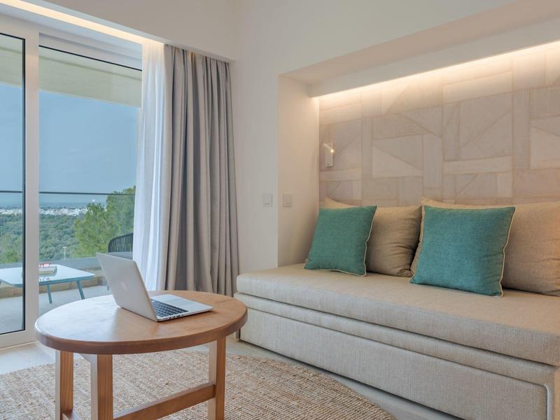 Area di stanza d'hotel moderna con divano, cuscini, tavolo rotondo in legno e balcone con vista sul verde.