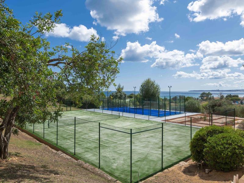 Campo sportivo all'aperto con campo da tennis, circondato da alberi e vista mare in una giornata soleggiata.
