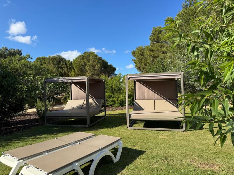 Comode lettini con tettucci in un prato circondato da alberi sotto un cielo blu.