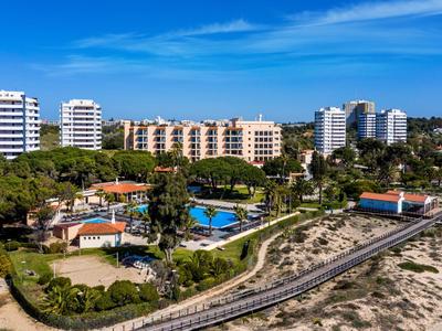 Hotelcomplex met zwembad omgeven door bomen nabij een strand en zandduinen onder een blauwe lucht.