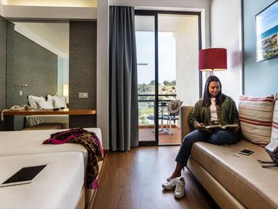 Vrouw zit op een bank in een moderne hotelkamer met bed en groot raam