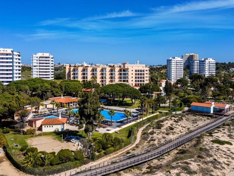 Hotelcomplex met zwembad omgeven door bomen nabij een strand en zandduinen onder een blauwe lucht.