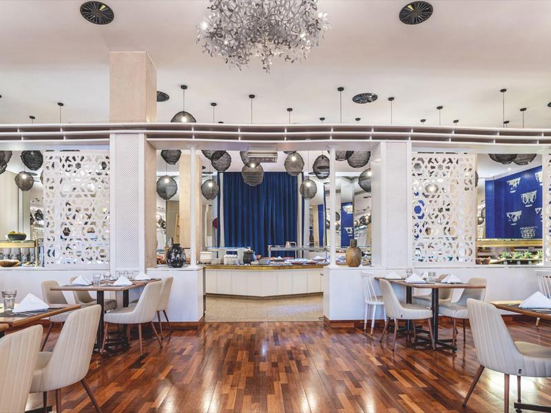 Elegant restaurant met houten vloer, moderne witte meubels en decoratieve kroonluchter.