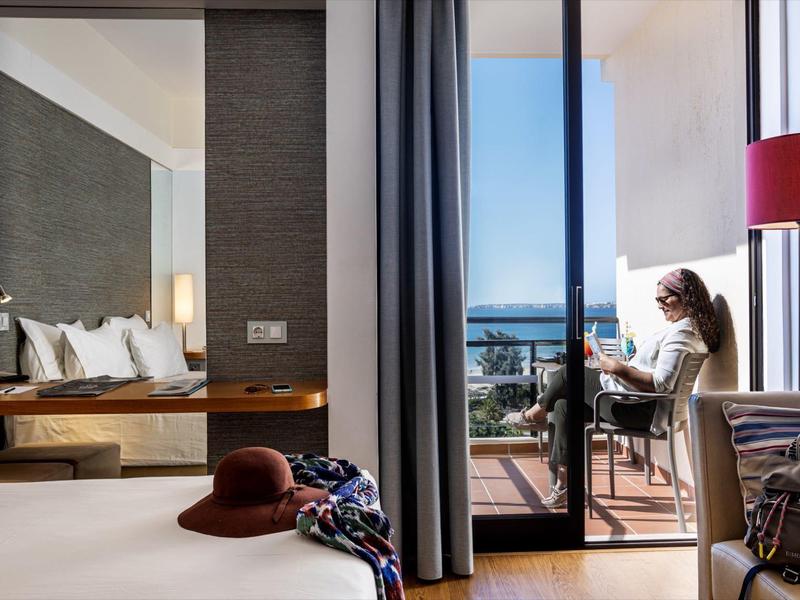 Hotelkamer met balkon, vrouw leest bij daglicht, uitzicht op stad en zee