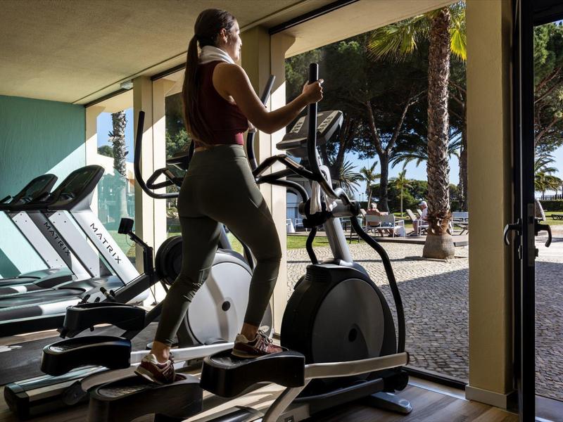Vrouw traint op crosstrainer in fitnessruimte met uitzicht op zonnig buitengebied.