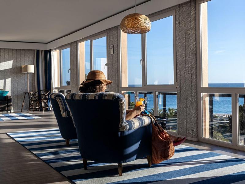 Hotel lounge met grote ramen, fauteuils en uitzicht op de zee op een zonnige dag.