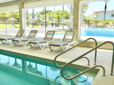Hallenbad mit Pooltreppe und Liegestühlen, große Fenster bieten Ausblick auf Außenpool und Garten.