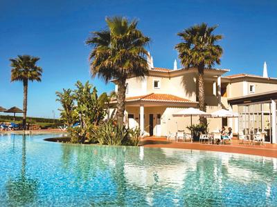 Großer Pool mit Palmen vor einem hellen Hotelgebäude an einem sonnigen Tag.