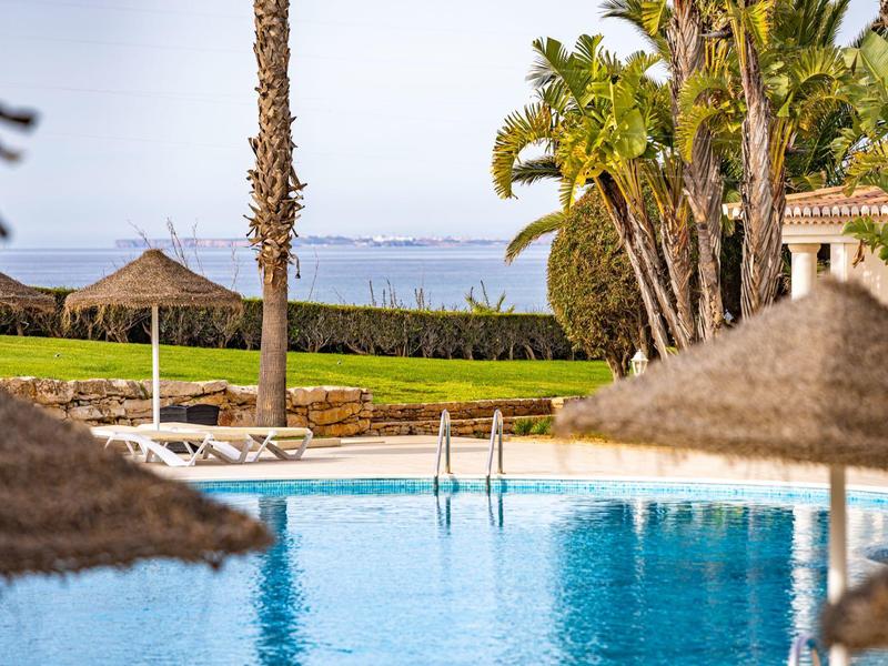 Ein blauer Pool mit Sonnenschirmen, Palmen und Meer im Hintergrund an einem sonnigen Tag.