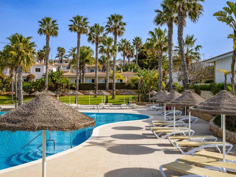 Poolbereich mit Palmen, Sonnenschirmen aus Stroh und Liegestühlen an sonnigem Tag